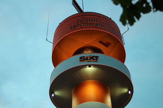 Das Sixt-Logo und der Slogan sowie das stilisierte Leuchtfeuer werden von der Brstung aus hinterleuchtet. Weitere Strahler und LED-Leuchtbnder illuminieren den Turm in der Dunkelheit